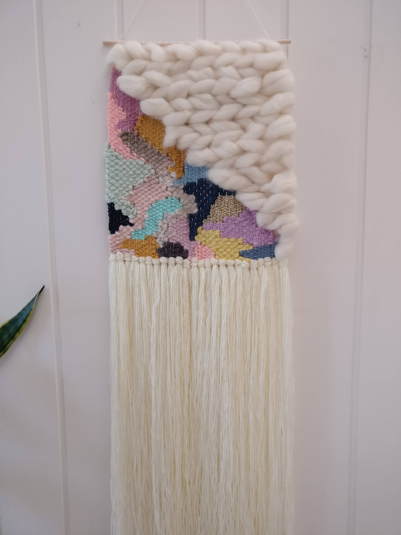 May include: A colourful woven wall hanging with a white fluffy texture at the top. The hanging has a fringe of white yarn at the bottom.
