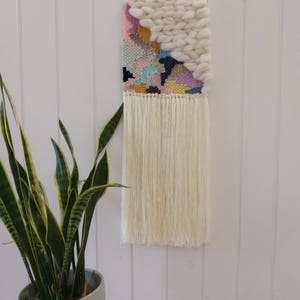 May include: A colourful woven wall hanging with a white fluffy texture at the top. The hanging has a fringe of white yarn at the bottom.