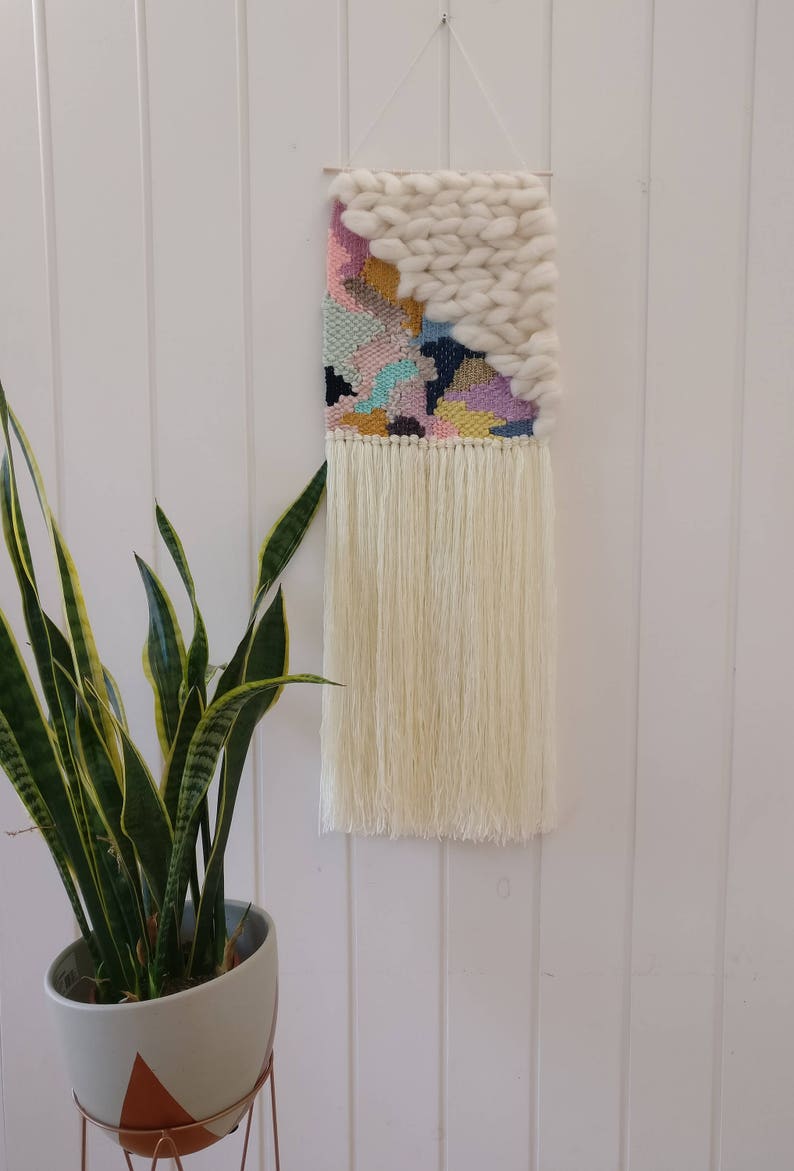 May include: A colourful woven wall hanging with a white fluffy texture at the top. The hanging has a fringe of white yarn at the bottom.