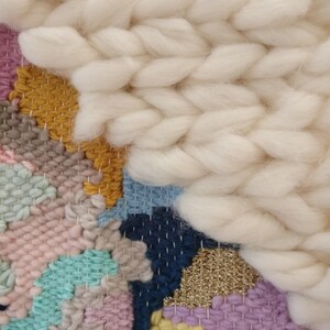 May include: A colourful woven wall hanging with a variety of textures and colours, including pink, blue, yellow, and white. The hanging features a large, fluffy white section in the top right corner.