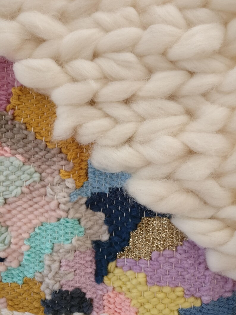 May include: A colourful woven wall hanging with a variety of textures and colours, including pink, blue, yellow, and white. The hanging features a large, fluffy white section in the top right corner.