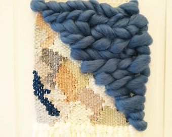Puede incluir: Un tapiz de pared tejido con un patrón geométrico en tonos beige, gris y blanco. La esquina superior derecha presenta un gran acento de lana trenzada azul.