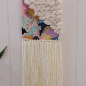 May include: A colourful woven wall hanging with a white fringe. The hanging features a geometric pattern with pink, blue, yellow, and green colours. The top of the hanging is made of a thick white yarn.