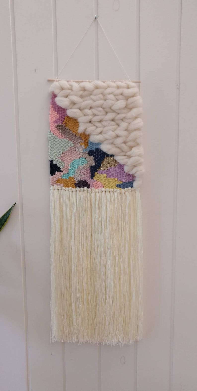 May include: A colourful woven wall hanging with a white fringe. The hanging features a geometric pattern with pink, blue, yellow, and green colours. The top of the hanging is made of a thick white yarn.