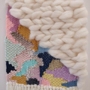 May include: A colourful woven wall hanging with a white fluffy texture on the top right corner. The hanging has a fringe border and features a variety of colours including pink, blue, yellow, and green.
