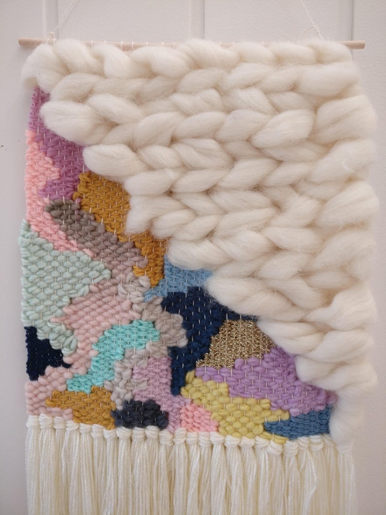 May include: A colourful woven wall hanging with a white fluffy texture on the top right corner. The hanging has a fringe border and features a variety of colours including pink, blue, yellow, and green.