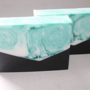 May include: Two bars of handmade soap with a swirling green and white design. The soap is topped with a black layer.