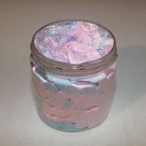 May include: A glass jar filled with a pink and blue whipped body butter with glitter on top.