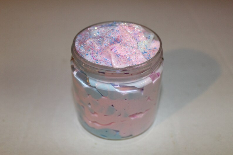 May include: A glass jar filled with a pink and blue whipped body butter with glitter on top.