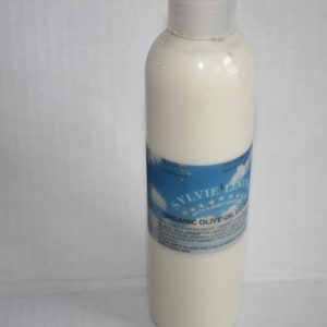 Puede incluir: Una botella de pl&aacute;stico blanca de loci&oacute;n de aceite de oliva org&aacute;nico con una etiqueta azul que dice "Sylvie Lind" y "Loci&oacute;n de aceite de oliva org&aacute;nico".