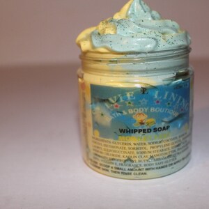 May include: A jar of whipped soap with a blue and yellow swirl on top. The jar has a label that says "Whipped Soap" and "The Silver Lining Bath & Body Boutique".