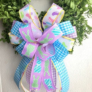 Può includere: Una ghirlanda pasquale pastello con un fiocco fatto di più nastri. I nastri presentano un motivo a quadri blu chiaro, un nastro viola con silhouette di conigli verdi e un nastro bianco con disegni di caramelle gommose gialle.