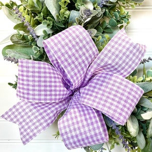 Può includere: Primo piano di una ghirlanda con un grande fiocco a quadretti vichy lavanda e bianco. La ghirlanda è composta da fogliame verde, tra cui orecchie d'agnello e rametti di lavanda. Lo sfondo è una superficie bianca a doghe.