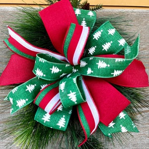 Puede incluir: Una corona de Navidad con un gran lazo rojo y verde. El lazo está hecho de cinta con una raya blanca y verde y una cinta verde con un patrón de árbol de Navidad blanco. La corona está hecha de ramas de pino.