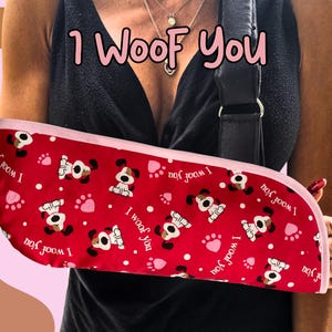 May include: Red arm sling featuring a pattern of cartoon dogs, paw prints, and the phrase "I woof you." The sling has a pink trim and is worn on the arm. The text "1 Woof You" is written across the top.