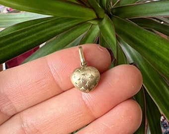 May include: A gold-colored heart-shaped pendant with a textured surface and starburst designs. The pendant is suspended from a small loop, ready to be added to a necklace or bracelet. The pendant is held in a hand, with a green plant in the background.