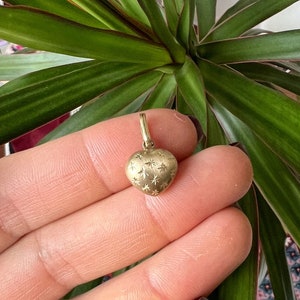 May include: A gold heart pendant with a textured finish and starburst patterns. The pendant hangs from a small loop. The pendant is held in a hand, with a green plant in the background. The pendant is designed to be added to a necklace or bracelet.