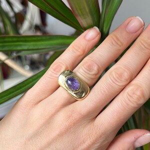 May include: A gold ring with a purple gemstone set in a rectangular bezel. The ring is worn on a finger.