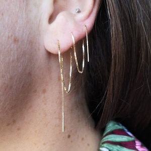 Puede incluir: Primer plano de la oreja de una persona con tres pendientes de cadena de oro. Los pendientes son de diferentes longitudes y estilos. El pendiente más largo es una cadena fina con una pequeña barra de oro en el extremo. El pendiente del medio es una cadena más gruesa con un pequeño aro de oro en el extremo. El pendiente más corto es un pequeño aro de oro.