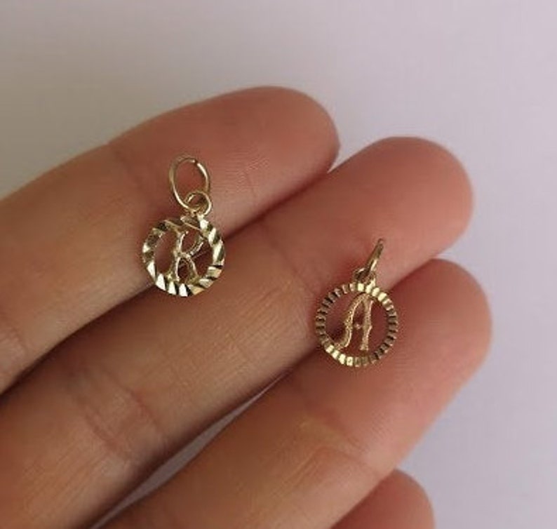 May include: Two gold-colored initial charms, one with the letter "K" and the other with the letter "A", both in a circular design with a textured edge.