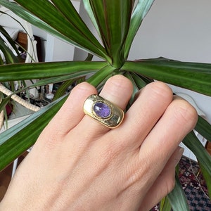 May include: A gold ring with a purple gemstone set in a textured oval bezel. The ring is thick and has a wide band.