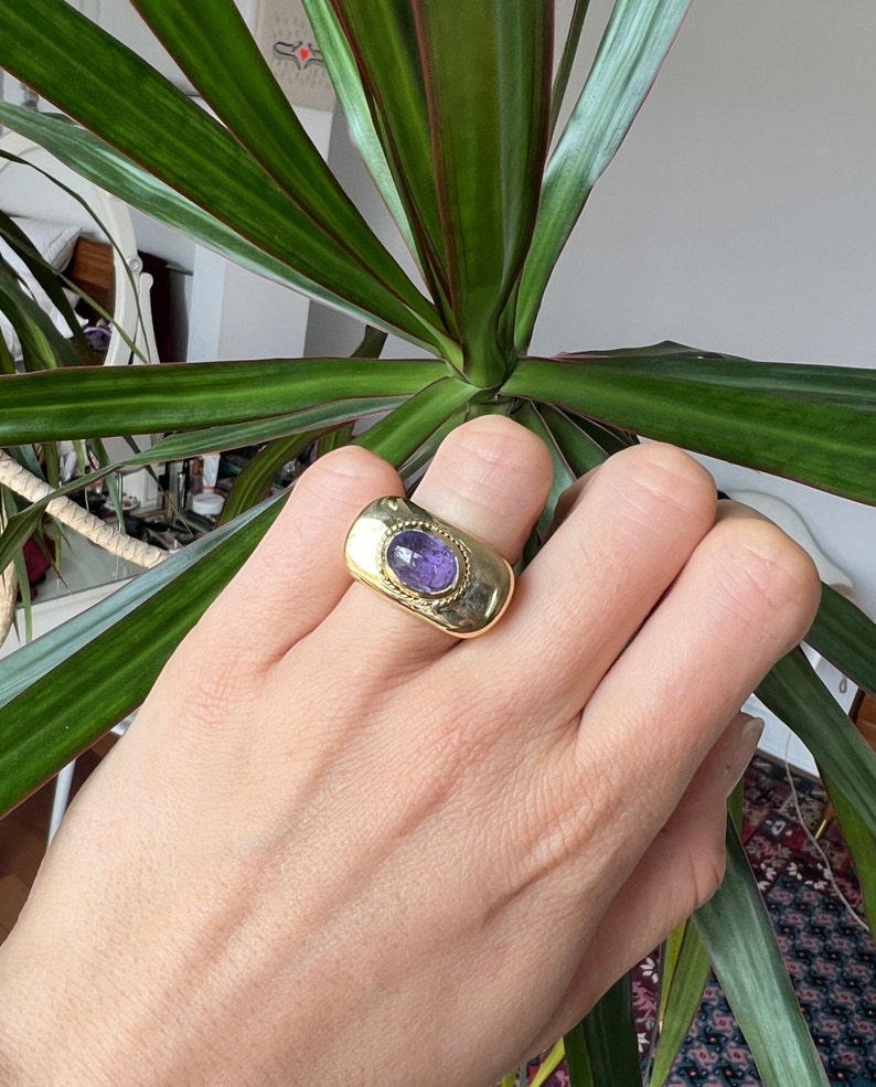 May include: A gold ring with a purple gemstone set in a textured oval bezel. The ring is thick and has a wide band.