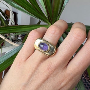 May include: A gold ring with a purple gemstone set in a rectangular bezel. The ring has a textured band.