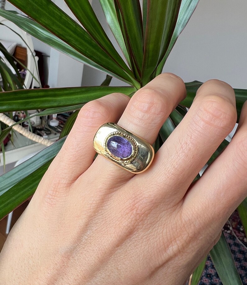 May include: A gold ring with a purple gemstone set in a rectangular bezel. The ring has a textured band.