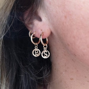 May include: Gold hoop earrings with a small charm dangling from each hoop. The charms are round and have the letters "B" and "S" embossed on them.