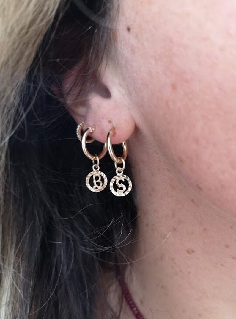 May include: Gold hoop earrings with a small charm dangling from each hoop. The charms are round and have the letters "B" and "S" embossed on them.