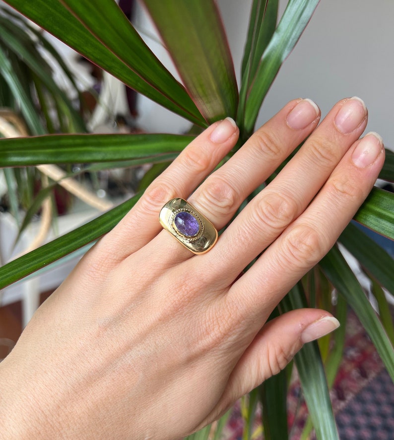 May include: A gold ring with a purple gemstone set in a rectangular bezel. The ring is worn on a finger.