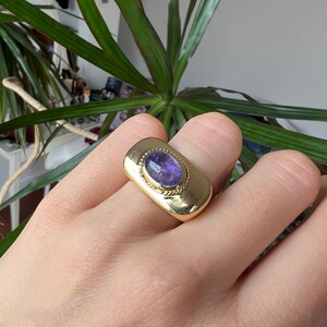 May include: A gold ring with a purple gemstone set in a rectangular bezel. The ring has a textured band.
