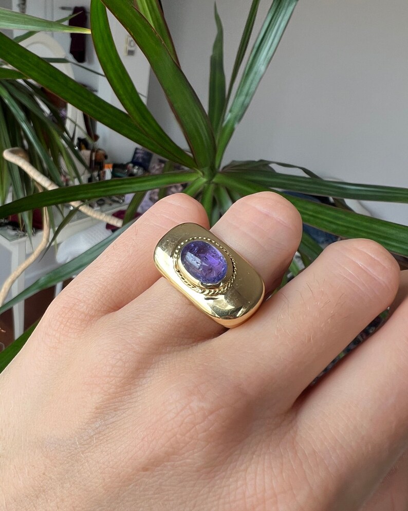 May include: A gold ring with a purple gemstone set in a rectangular bezel. The ring has a textured band.
