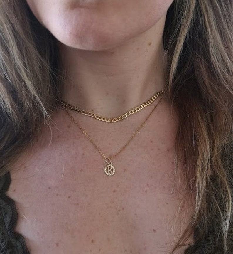 May include: A gold chain necklace with a small pendant featuring the letter K. The necklace is layered over a thicker gold chain.
