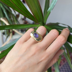 May include: A gold ring with a purple gemstone set in a rectangular bezel. The ring has a textured band.