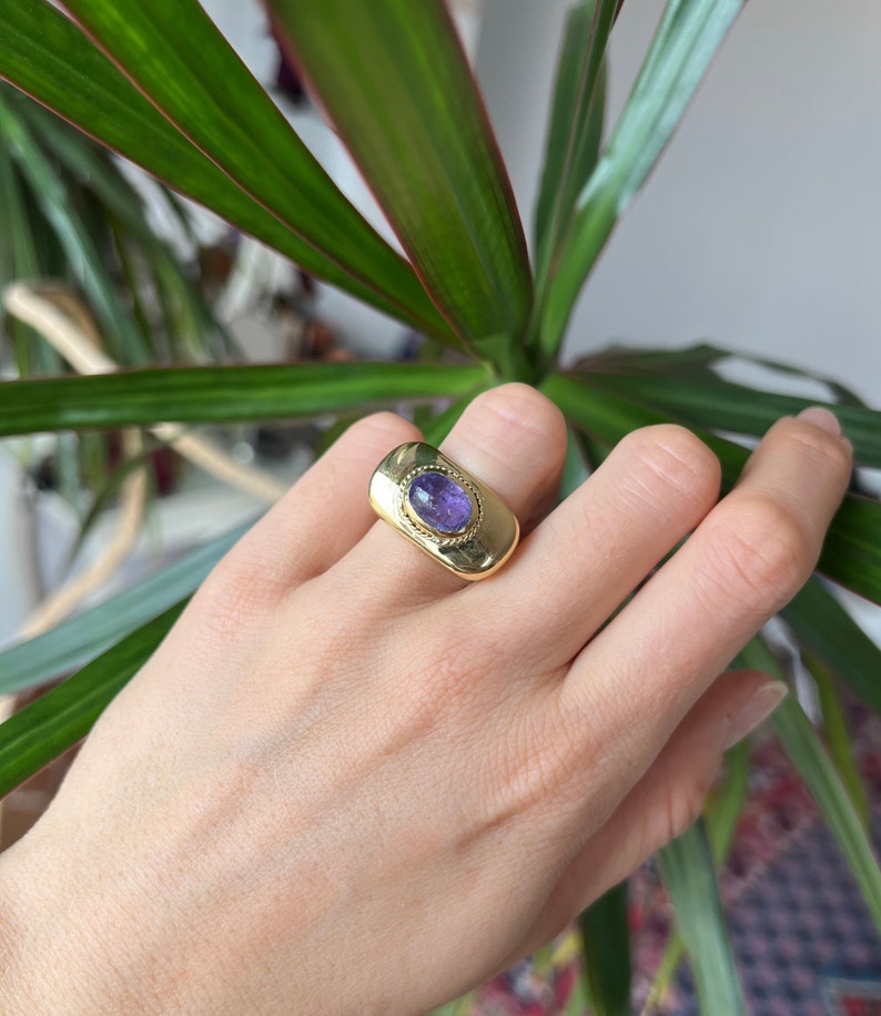 May include: A gold ring with a purple gemstone set in a rectangular bezel. The ring has a textured band.