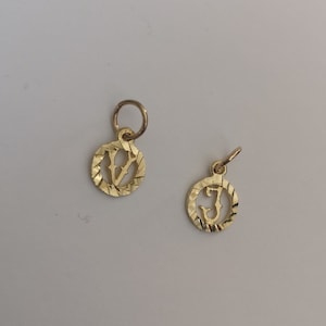 May include: Two gold-colored charms with a textured circular design. The charms feature the letters "V" and "J" in a stylized font.