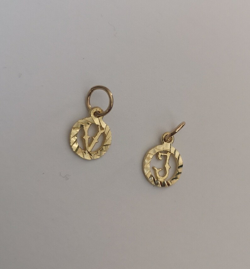 May include: Two gold-colored charms with a textured circular design. The charms feature the letters "V" and "J" in a stylized font.