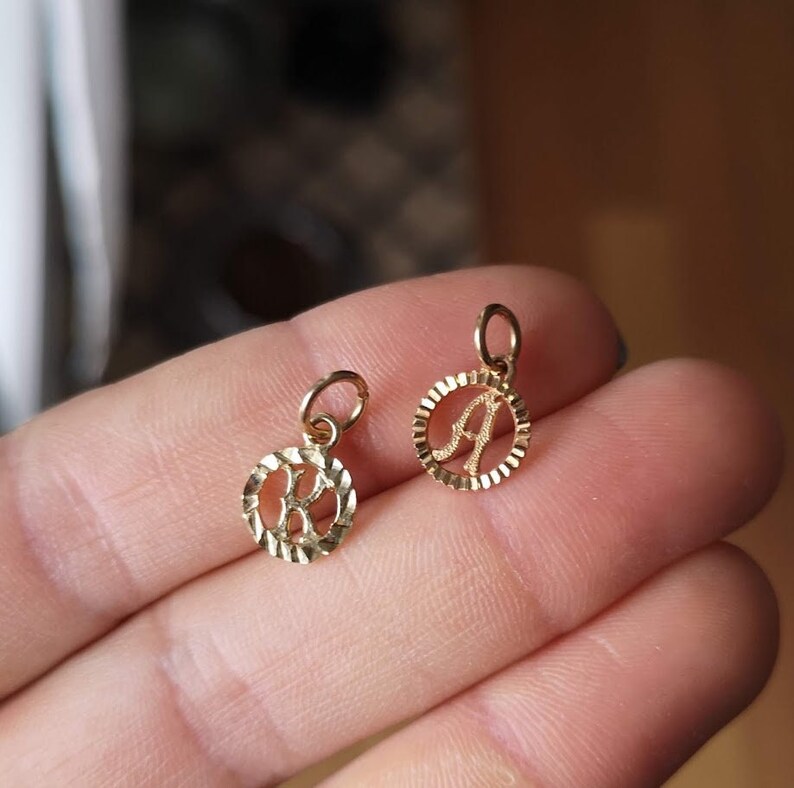 May include: Two gold-colored initial charms, one with the letter 'A' and the other with the letter 'K', both with a textured circular design.