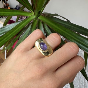 May include: A gold ring with a purple gemstone set in a rectangular bezel. The ring has a textured band.