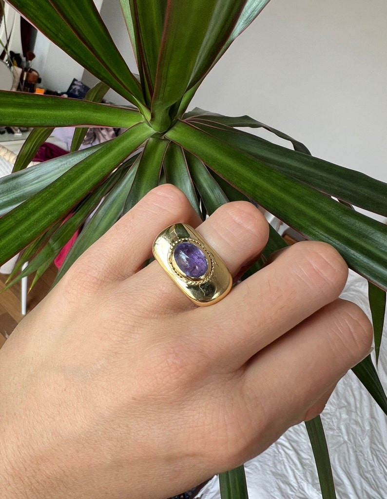 May include: A gold ring with a purple gemstone set in a rectangular bezel. The ring has a textured band.