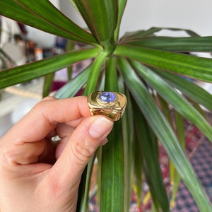 May include: A gold ring with a large, oval-shaped, purple gemstone set in a gold bezel. The ring has a textured band.
