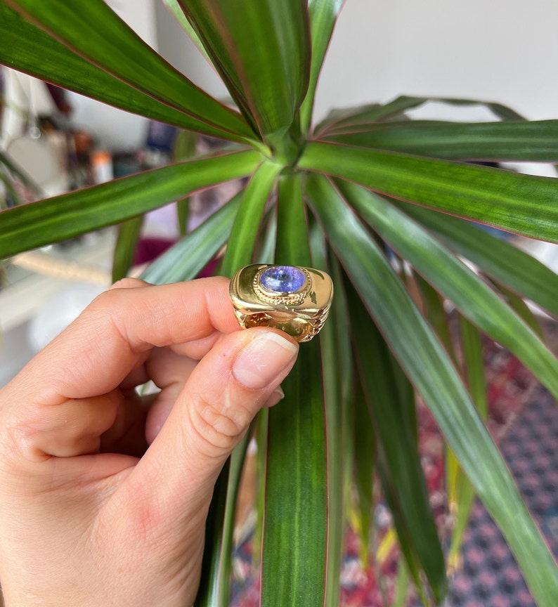May include: A gold ring with a large, oval-shaped, purple gemstone set in a gold bezel. The ring has a textured band.