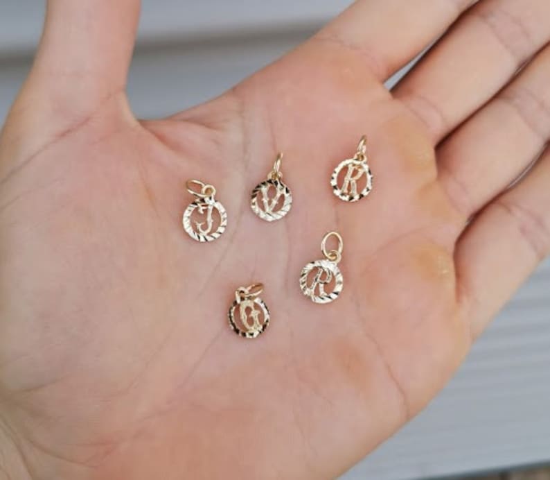 May include: Five gold-colored initial charms with a textured circular design. The charms are for the letters J, V, R, R, and G.