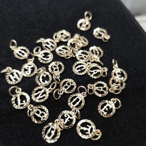 May include: A collection of gold-colored alphabet letter charms. Each charm is a circle with a raised letter in the center. The charms are scattered on a black surface.