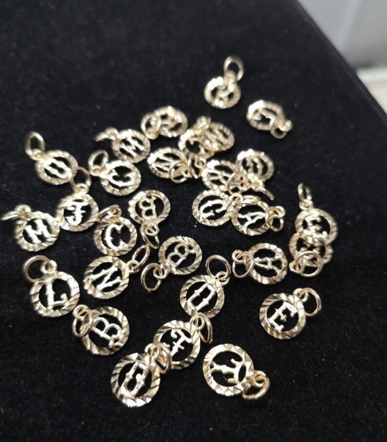 May include: A collection of gold-colored alphabet letter charms. Each charm is a circle with a raised letter in the center. The charms are scattered on a black surface.