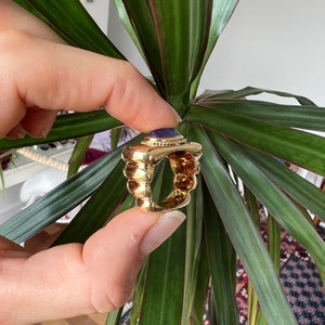 May include: A gold ring with a large, round, purple gemstone set in the center. The ring has a textured, ridged band.