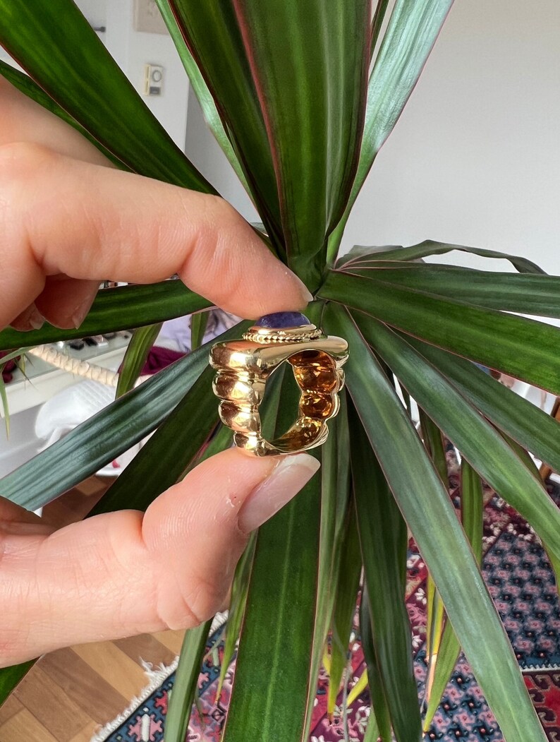 May include: A gold ring with a large, round, purple gemstone set in the center. The ring has a textured, ridged band.