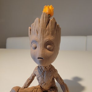 Puede incluir: Una figurita de Groot, personaje de la película Guardianes de la Galaxia, impresa en 3D en color marrón, sentada con una pequeña planta naranja en la cabeza.