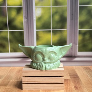 May include: A light green planter shaped like a small creature with large pointed ears and wide eyes. The planter features a circular opening at the top and a textured collar detail around the base, resting on a small wooden crate structure.